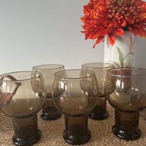 May include: A set of five matching drinking glasses made of translucent brown glass. Each glass features a rounded bowl shape that tapers down into a cylindrical stem and a circular base. The glasses are arranged in a group on a textured surface.