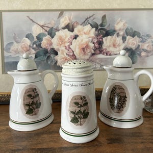 May include: A set of three white ceramic kitchen containers featuring green speckled lids and botanical illustrations. Two pitchers have handles and lids, while the central cylindrical shaker has a perforated top and the text Cheese.