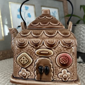 May include: A brown ceramic teapot shaped like a house with a black wire handle. The exterior features embossed scalloped roof details, a central door with two black windows, and various geometric and floral relief decorations on the front facade.