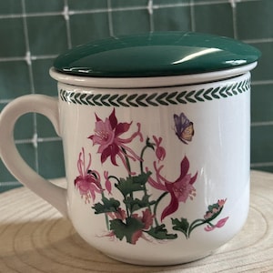 May include: A white ceramic mug with a dark green lid, featuring a botanical illustration of pink columbine flowers and a small butterfly. The rim is decorated with a repeating green leaf pattern, and the mug has a single curved handle.