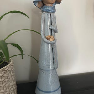 May include: A tall, slender ceramic figurine with a speckled light blue glaze. The figure wears a wide-brimmed hat and a long coat, holding a small cluster of dried beige flowers in its hands. The base is flared and features a horizontal blue line.