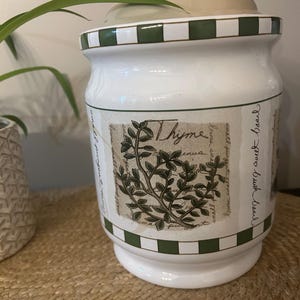 May include: A white ceramic canister with a lid, featuring a green and white chequered border at the top and bottom. The centre displays a botanical illustration of a thyme plant with the word Thyme and cursive script text on a beige background.