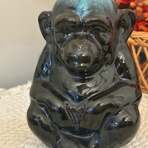 Peut inclure: Une figurine de singe en céramique brillante avec un corps brun foncé à la finition lisse et réfléchissante, contrastée par un émail bleu sarcelle sur le sommet de la tête. Le singe est représenté en position assise, les bras sur les genoux.