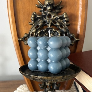 May include: A square-shaped bubble candle in a muted slate blue colour, featuring a grid of spherical wax spheres and a single white wick centred on top. The candle is positioned on a dark, weathered metal shelf with a decorative, ornate backplate.