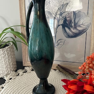 May include: A tall, slender ceramic pitcher with a glossy finish, featuring a deep teal and black gradient glaze. The vessel has a curved handle, a narrow neck, and a flared base, standing upright with a smooth, reflective surface texture.