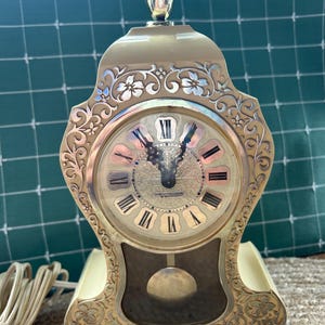 Peut inclure: Une horloge de cheminée électrique Westclox couleur crème avec un fleuron doré et des volutes florales ornées. Le cadran présente des chiffres romains noirs, des aiguilles décoratives et un pendule central visible par une vitre.