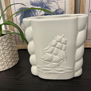 Peut inclure: Un vase en céramique blanche avec une finition brillante, présentant un relief moulé d'un voilier sur le devant. Le vase possède un design distinctif à bords verticaux festonnés ou arrondis des deux côtés, créant un aspect texturé.