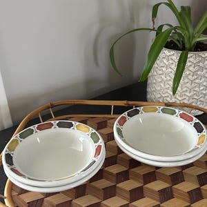 Peut inclure: Deux piles de bols en céramique blanche avec un motif décoratif sur le bord. Le bord présente des segments rectangulaires alternés dans des tons de vert, jaune, marron et rouge, séparés par de petites icônes en forme d'os sur fond sombre.