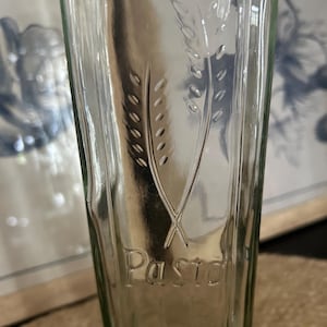 Peut inclure: Un récipient de stockage en verre transparent avec un profil carré, présentant un motif en relief de deux épis de blé et le mot "Pasta" imprimé en lettres surélevées sur la surface avant. Le verre a une finition lisse et transparente.