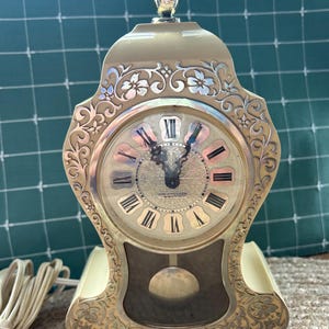 Peut inclure: Une horloge de cheminée électrique Westclox couleur crème avec un fleuron doré, des volutes florales argentées ornées et un cadran rond avec des chiffres romains noirs. L'horloge comprend un pendule visible derrière une vitre et un cordon.