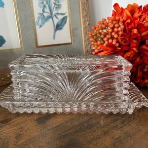 May include: A clear pressed glass butter dish featuring a rectangular base and a matching lid. The glass surface is decorated with a repeating fan-like pattern of raised ridges and a textured, scalloped edge along the bottom of the lid and base.