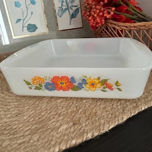 May include: A square white milk glass baking dish featuring a colourful floral pattern along the front side. The design includes orange, yellow, and blue flowers with green leaves and stems. The dish has integrated handles on two sides.