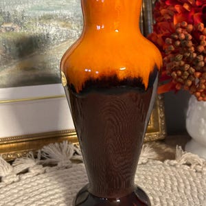 Peut inclure: Un vase en céramique à la finition brillante, présentant une partie supérieure orange vif qui se fond dans une base brun foncé. Le corps inférieur du vase affiche une texture subtile de grain de bois vertical, avec un bord lisse et arrondi.