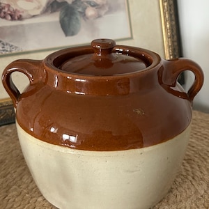 May include: A two-handled ceramic crock with a lid, featuring a glossy brown glazed upper section and a matte cream-coloured lower base. The vessel has a rounded body, two side handles, and a small knob on the centre of the circular lid.