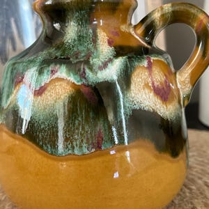 May include: A ceramic pitcher with a rounded body and a single handle. The glaze features a glossy finish with a mustard yellow base, transitioning into a drip pattern of forest green, dark brown, and hints of deep burgundy near the rim.