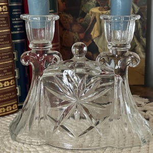 May include: A clear glass dual candle holder featuring a central lidded compartment with a starburst pattern. The piece includes two vertical candle sockets, each holding a light blue twisted taper candle, and a small round knob on the lid.