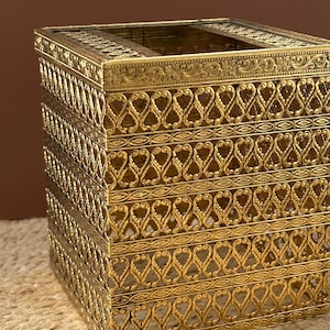 May include: A square tissue box cover made of gold-toned metal with an intricate filigree design. The exterior features repeating openwork patterns of vertical loops and horizontal bands with embossed floral details along the top edge.
