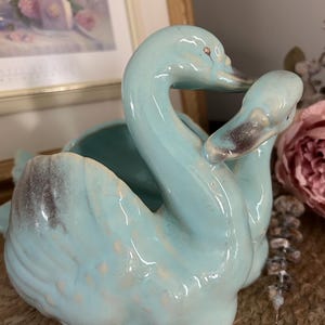 Peut inclure: Un cache-pot en céramique bleu clair représentant deux cygnes aux cous courbés l'un vers l'autre. La finition brillante présente des nuances grises subtiles sur les ailes et les becs, avec un centre creux pour contenir des objets.