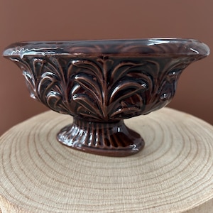 May include: A dark brown ceramic bowl with a glossy finish, featuring a raised leaf pattern around the exterior and a pedestal base with vertical ridges. The bowl has a wide, flared rim and a deep, rounded interior basin.