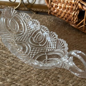 May include: A clear pressed glass dish with an elongated oval shape and a single handle. The surface features a detailed pattern of geometric cross-hatching, fan-like motifs, and curved lines, with a scalloped edge along the entire rim.