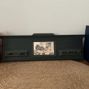 May include: A rectangular black wall-mounted rack featuring a central inset tile illustration of a hen with chicks near a coop. The rack includes two horizontal sections on either side, each equipped with two small black knobs for hanging items.