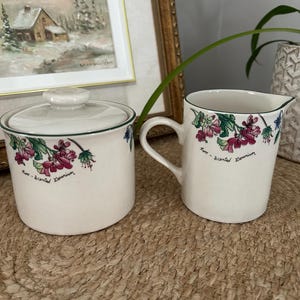 Peut inclure: Un sucrier en céramique crème avec couvercle et un pot à lait assorti. Les deux pièces présentent une illustration botanique de fleurs de géranium rose odorant et de feuilles vertes, avec le texte Rose-Scented Geranium écrit en noir.