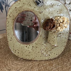 May include: A square-shaped ceramic wall mirror with rounded corners, featuring a speckled light green glaze. An oval mirror is embedded in the centre, and a small attached ceramic vase holds a dried brown flower and cream-coloured baby's breath.