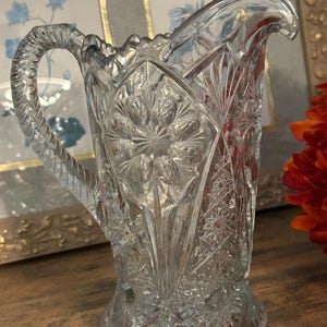 Peut inclure: Un pichet en verre transparent présentant un motif pressé complexe avec des étoiles, des tailles en diamant géométriques et un bord festonné. Le récipient comprend une anse robuste au design texturé en corde et une base large et stable.