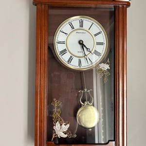 May include: A rectangular wall clock with a dark wood frame and a glass front. The white clock face features black Roman numerals, the text Masterpiece and Quartz, and black hands. A gold-toned pendulum hangs below, with floral designs on the glass.