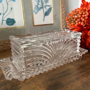 Peut inclure: Un beurrier en verre transparent composé d'une base rectangulaire et d'un couvercle assorti. Le couvercle présente un motif en éventail rayonnant depuis le centre, tandis que la base possède un bord festonné et dentelé sur tout le pourtour.