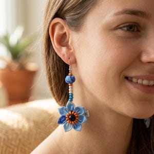 Könnte beinhalten: Ein Hängeohrring mit einer hellblauen Stoffblume, deren dunkle Mitte von kleinen orangefarbenen Perlen umgeben ist. Über der Blume befinden sich mehrere kleine Perlen in Blau, Weiß und Orange, gekrönt von einer blauen Stoffkugel an einem Silberhaken.