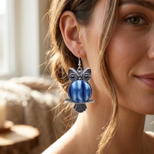 Könnte beinhalten: Ein silberfarbener Eulen-Ohrring mit einem runden, blau-weiß gemusterten Tie-Dye-Stoffeinsatz als Körper. Das Eulen-Design umfasst detaillierte Metallflügel, einen Schwanz und dunkle, runde Augen, befestigt an einem silberfarbenen Ohrhaken.