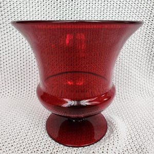 May include: A deep red glass vase with a wide, flared rim and a rounded body that tapers down to a circular pedestal base. The glass is translucent and features a smooth, polished surface, reflecting light across its curved, symmetrical form.