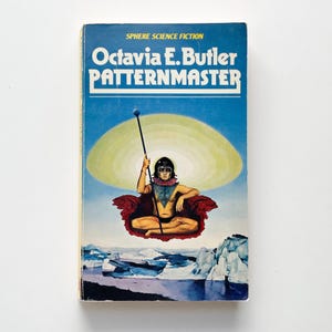 May include: A vintage paperback copy of Octavia E. Butler's "Patternmaster". The cover art depicts a science fiction scene with a figure holding a staff, set against a blue sky, a yellow halo, and an icy landscape.