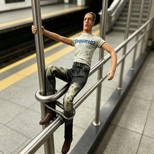 Peut inclure: Une figurine articulée au teint clair, portant un t-shirt gris à manches courtes avec le texte bleu DSQUARED2 imprimé sur la poitrine, associé à un pantalon en jean gris foncé délavé et des chaussures marron, positionnée sur une rampe en métal.