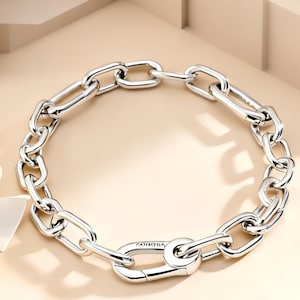 May include: A silver-toned chain link bracelet featuring elongated, polished oval links and a prominent, functional carabiner-style clasp closure. The metal surface reflects light, highlighting the smooth, rounded edges of each individual link.