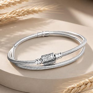 May include: A silver snake chain bracelet featuring a rectangular clasp encrusted with small clear stones. The bracelet has a flexible, woven metal texture and a polished finish, designed to be worn as a double-wrap piece around the wrist.