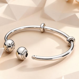 May include: A polished silver-tone open bangle bracelet featuring a smooth, circular design. The bracelet includes two spherical end caps, one of which is detachable, and two smaller fixed beads along the band. The word PANDORA is engraved on the beads.