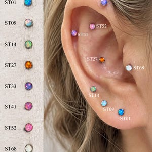 May include: A collection of round, cabochon-style stud earrings featuring various colors labeled ST01, ST09, ST14, ST27, ST33, ST41, ST52, and ST68. The studs are displayed in a vertical column and also shown individually inserted into multiple ear piercings.