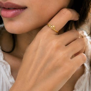 May include: A gold-toned ring featuring the name Aria in a cursive script font, with a small crown icon positioned above the letter i. The ring has a thin, polished band that encircles the finger, showcasing a metallic finish and detailed lettering.