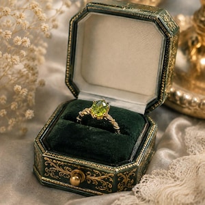 May include: A gold ring featuring a central oval-cut lime green gemstone held by prongs, with a band accented by small clear stones. The ring is displayed inside an octagonal dark green velvet-lined jewellery box with gold filigree detailing.