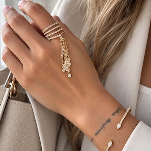 Op de afbeelding: Een goudkleurige ring met meerdere banden en een bungelend kettingkwastje met kleine ronde doorzichtige steentjes, gecombineerd met een bijpassende open armband met marquise-geslepen stenen aan beide uiteinden, afgewerkt in gepolijst goud.
