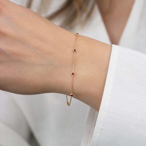 Puede incluir: Una delicada pulsera de cadena dorada con pequeñas piedras preciosas redondas de color rojo intenso, espaciadas uniformemente y engastadas en biseles dorados. La pulsera consiste en una fina cadena de eslabones que rodea la muñeca.