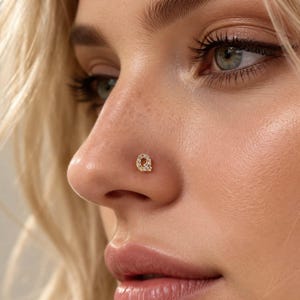 May include: A gold-toned nose stud shaped like the letter Q, featuring small, clear, sparkling stones embedded along the surface of the letter. The jewelry is positioned on the side of a nostril, showcasing a polished metallic finish and clear gems.