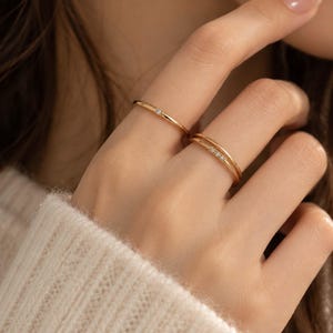Puede incluir: Dos anillos finos de oro en los dedos. Un anillo presenta una sola piedra pequeña y clara, mientras que el otro es un diseño de doble banda con tres piedras pequeñas y claras dispuestas en fila sobre la superficie superior de las bandas.