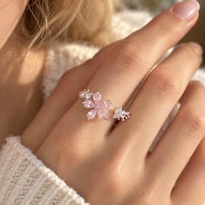 Puede incluir: Un anillo de tono plateado con un diseño floral central compuesto por cinco piedras de color rosa claro en forma de lágrima. La banda está decorada con pequeñas piedras transparentes y brillantes dispuestas en grupos a ambos lados de la flor.