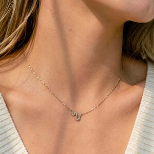 Puede incluir: Un collar de cadena en tono dorado con un pequeño colgante en forma de serpiente incrustado con diminutas piedras claras y brillantes. La delicada cadena de metal descansa sobre el cuello, mostrando el diseño detallado y curvo del dije.