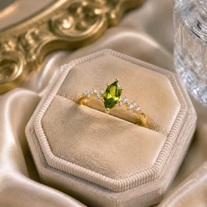 May include: A gold ring featuring a central marquise-cut olive green gemstone flanked by small clear stones on each side. The band is thin and polished, showcasing a delicate design that highlights the vibrant green stone and the sparkling accents.