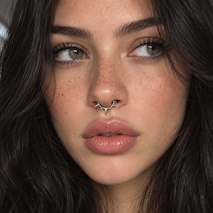 Puede incluir: Un anillo de septum de plata con un diseño de barra curva y cinco pequeñas púas espaciadas uniformemente que apuntan hacia abajo. La joya metálica está colocada en el centro de la nariz, mostrando un acabado pulido y un aspecto definido.