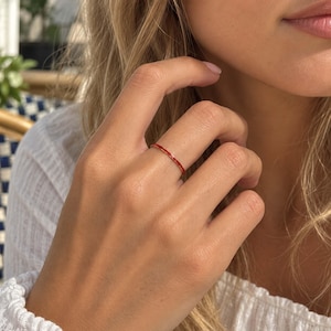 May include: A thin, minimalist ring featuring a vibrant red enamel finish on a smooth, slender band. The ring is worn on the finger, showcasing its simple, circular design and bright, solid red color that wraps entirely around the band.